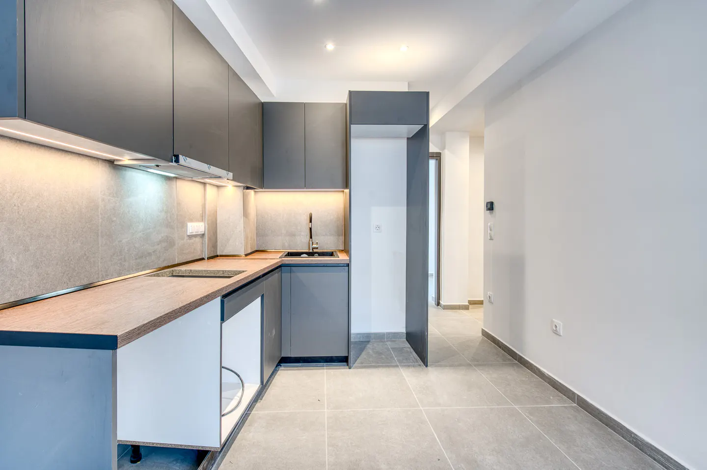 A modern kitchen with gray cabinets, wood countertops, and gray tile flooring. A hallway is visible in the background.