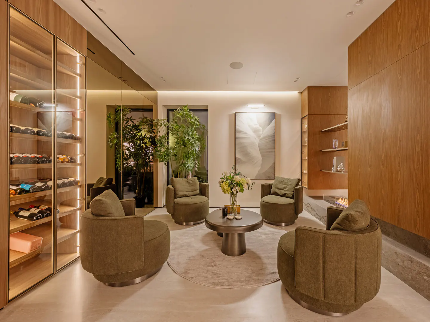 A modern living room with olive green chairs arranged around a round table on a light rug. A wine rack and fireplace are visible.