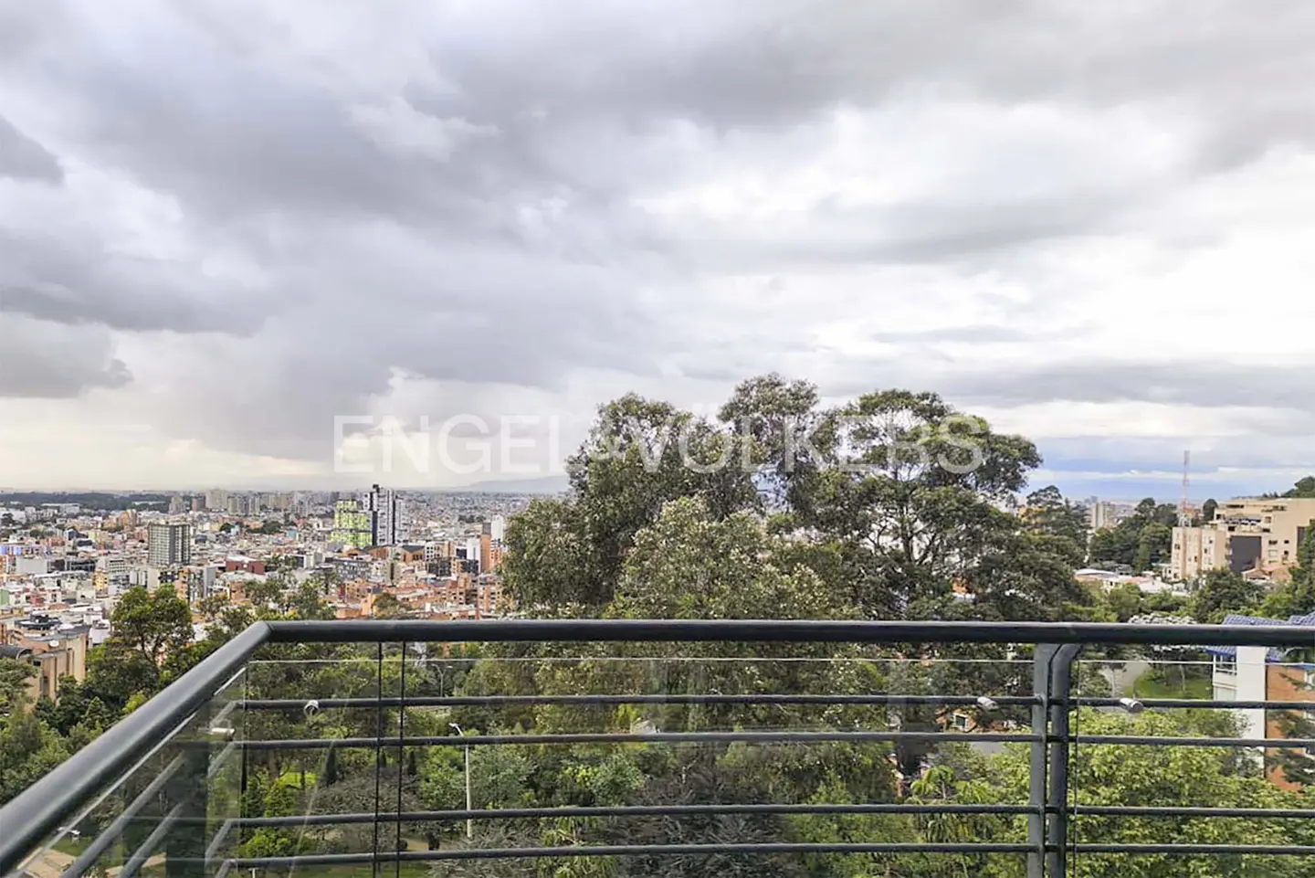 View from a balcony with a black metal railing overlooking trees and a city skyline under a cloudy sky.
