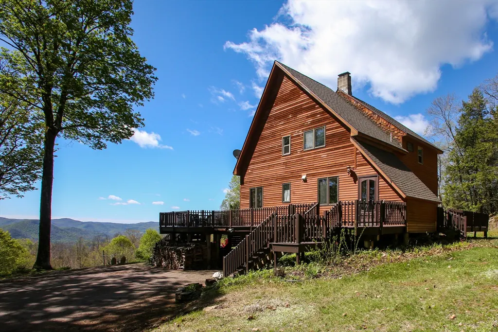 Exterior view of a wood-sided A-frame house with a deck, set on a grassy hill with a mountain view under a blue sky.