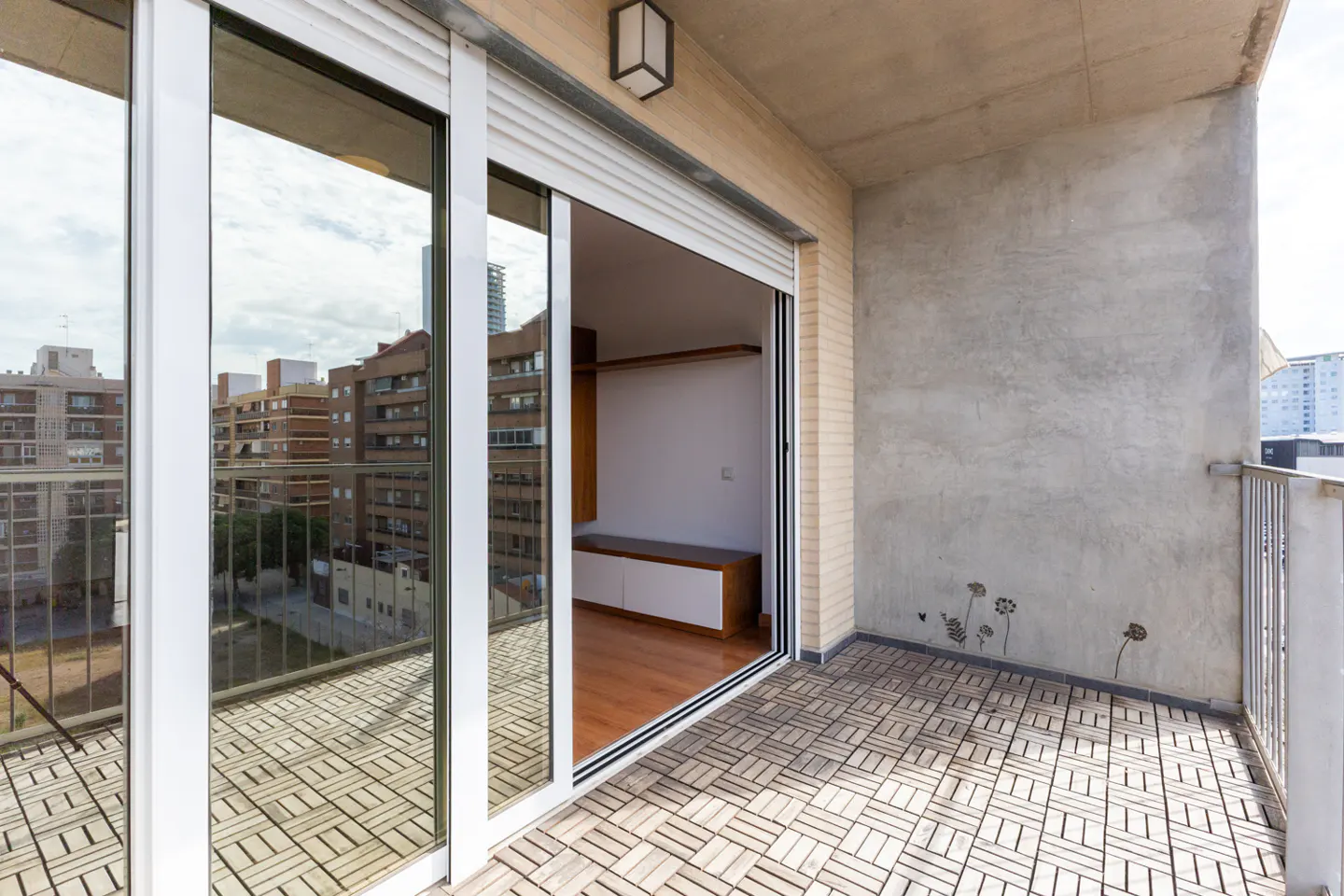 A balcony with a sliding glass door open to a room with wood floors and a white cabinet. The balcony has a gray tiled floor and a concrete wall.