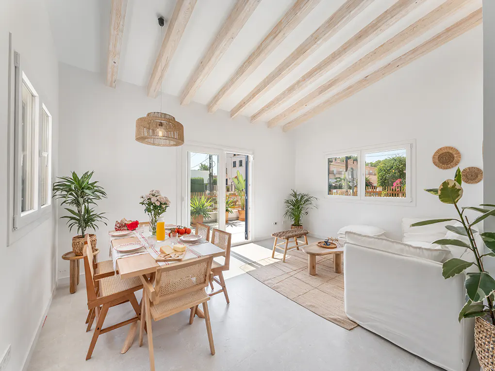 Bright, airy living space with white walls, exposed wood beams, and a dining table set for a meal. Sliding glass doors lead to a sunny patio.