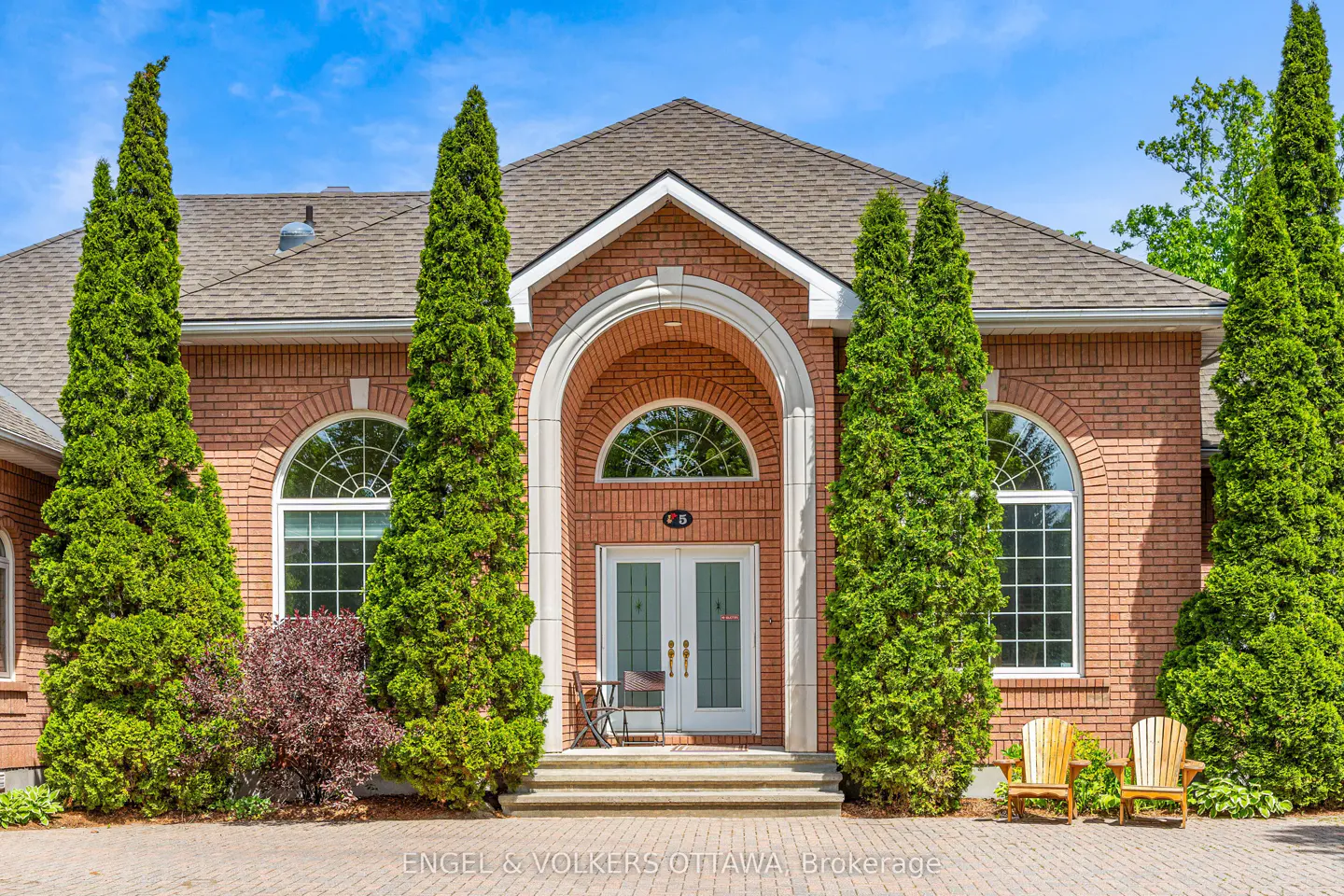 Front view of a red brick house with a white arched entryway, tall evergreen trees, and two wooden chairs on the patio.