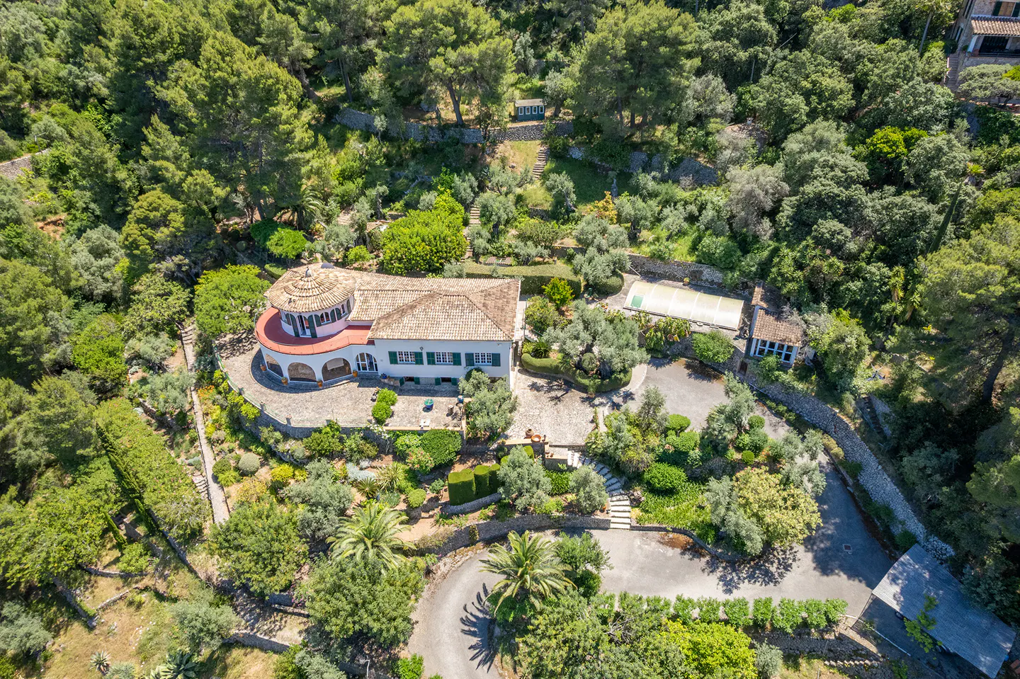 Aerial view of a white villa with a terracotta roof, surrounded by lush green trees, gardens, and a winding driveway.