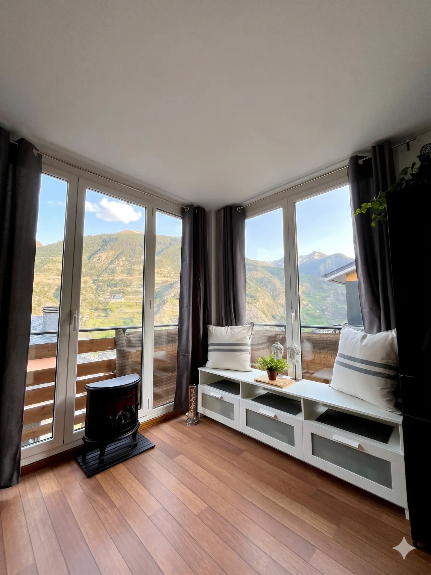 Bright room with wood floors, large windows, and mountain views. A black stove sits near the windows, and a white cabinet with pillows is against the wall.