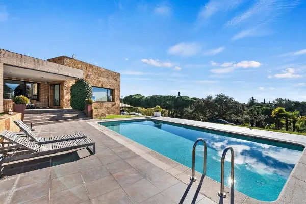 Exterior view of a stone house with a blue swimming pool, lounge chairs, and a clear blue sky.