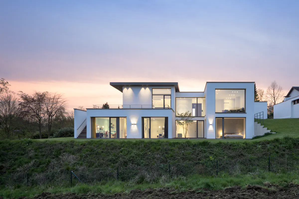 Modern white house with large windows on a green hill under a pink and blue sky.