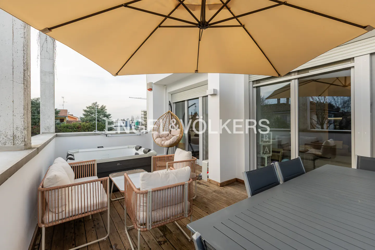 A balcony with a hot tub, chairs, a table, and a large umbrella. The balcony has a wooden floor and a view of the city.