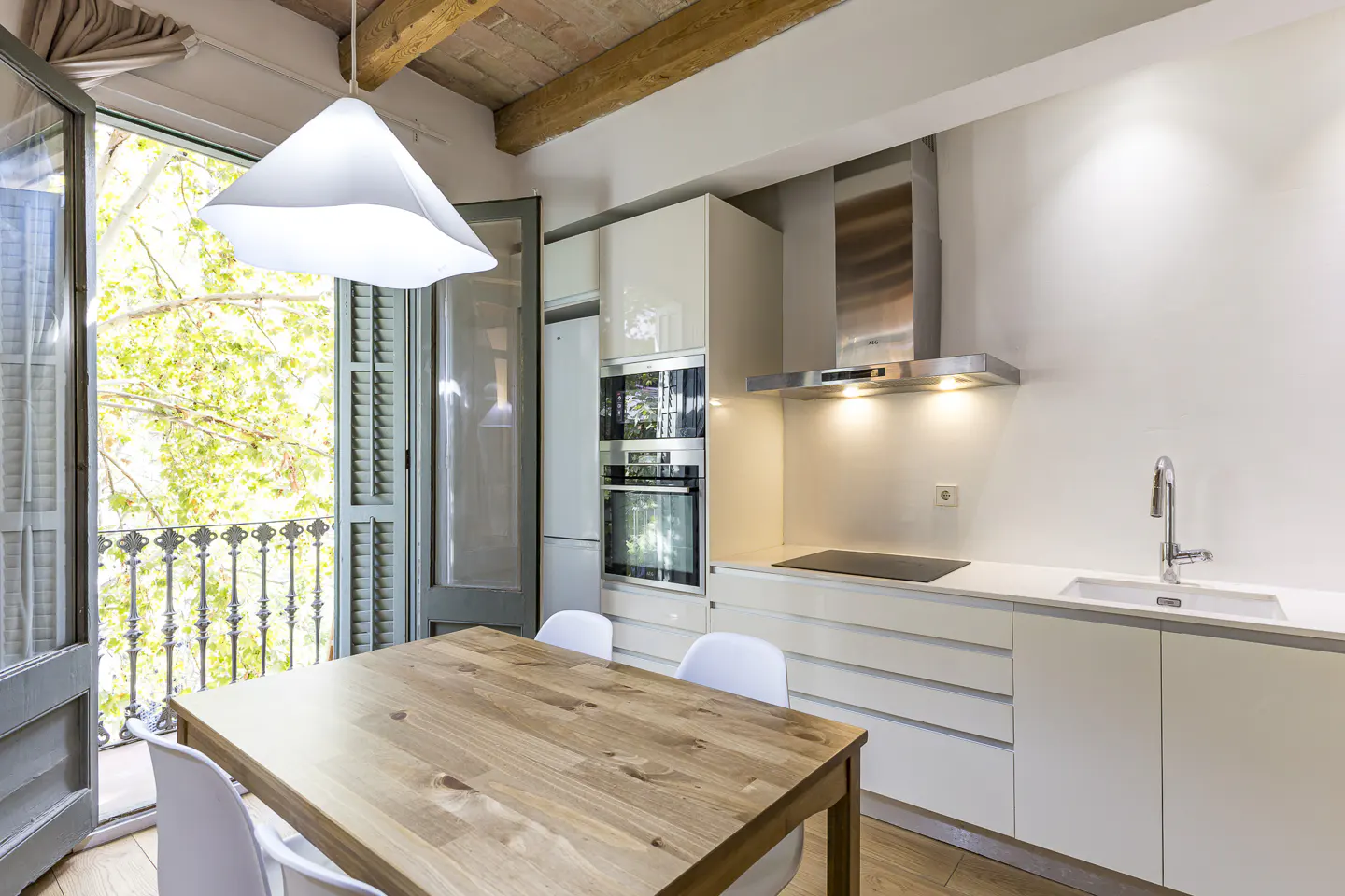 Bright kitchen with a wooden table, white chairs, and a balcony with green trees visible through the open doors.