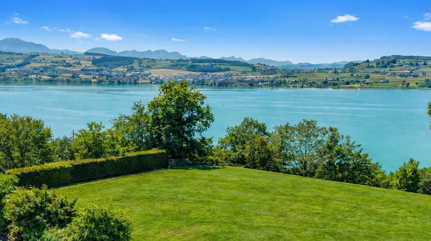 Scenic view of a turquoise lake, green lawn, trees, and distant mountains under a blue sky.