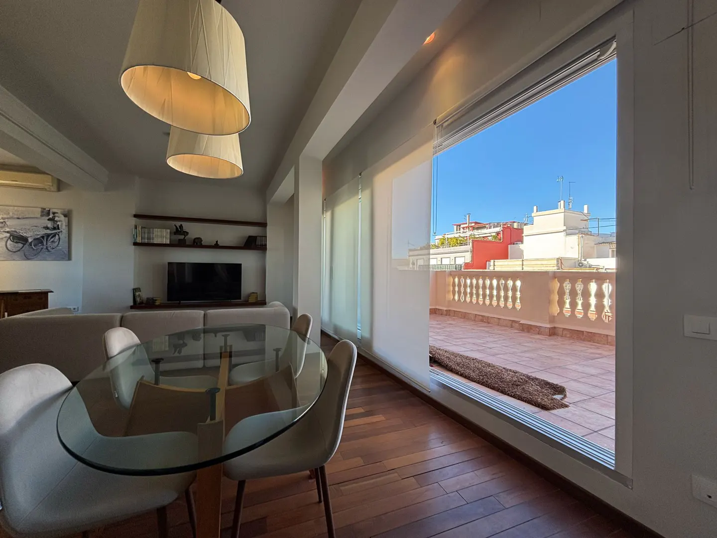Bright living room with a glass table, chairs, and a large window showing a rooftop terrace with city views.