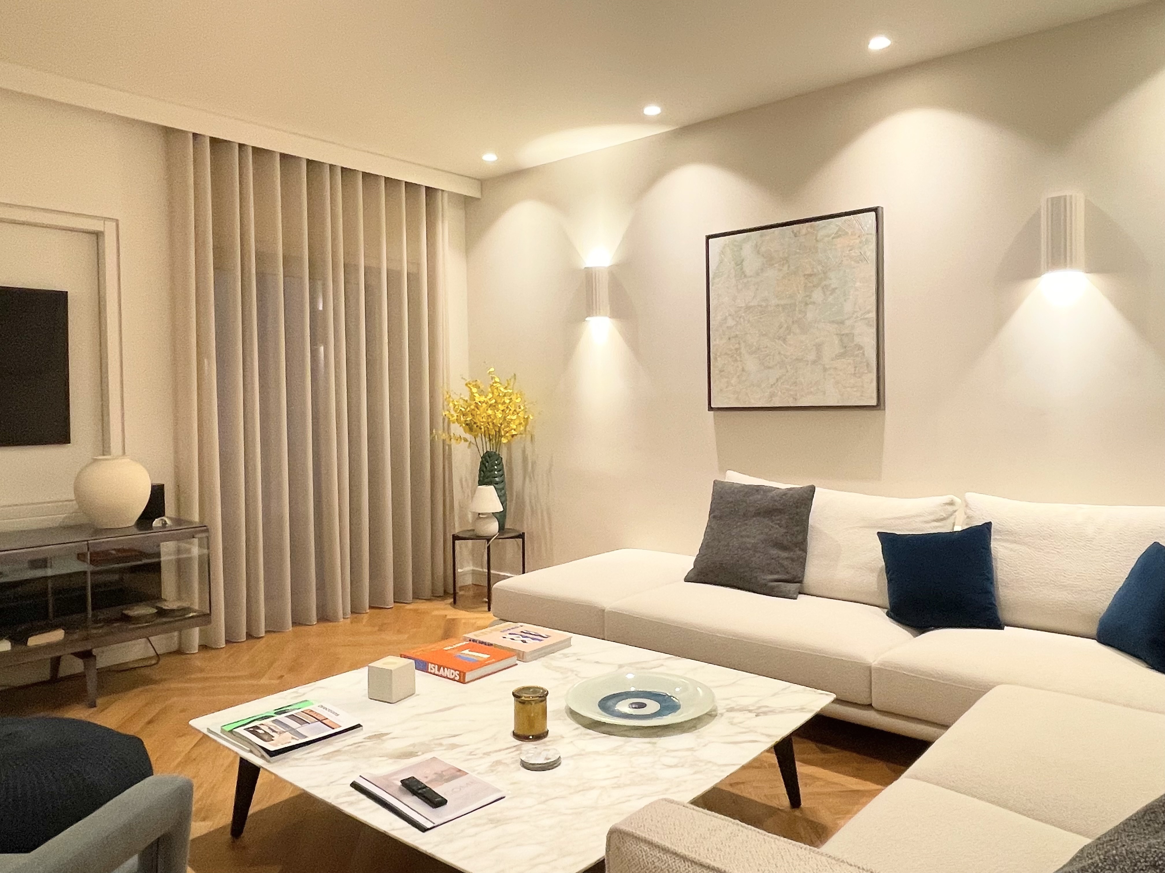 Modern living room with a white sectional sofa, marble coffee table, floor-to-ceiling curtains, and warm wall lighting. [AI-generated text]