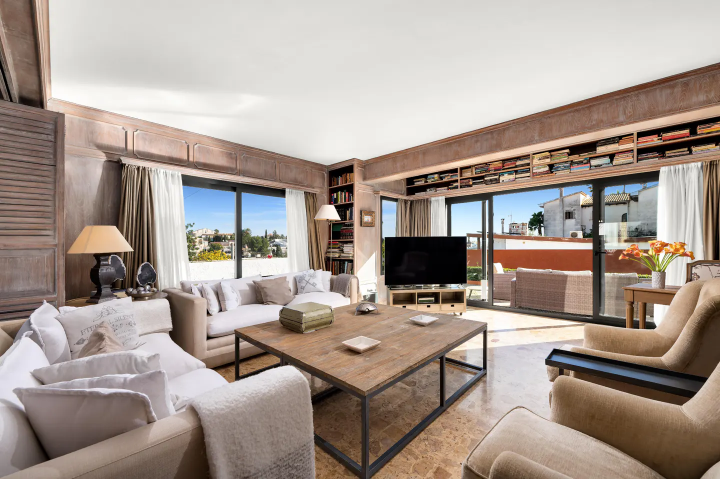 Bright living room with sofas, chairs, and a wood coffee table. Bookshelves line the walls above sliding glass doors to a balcony.