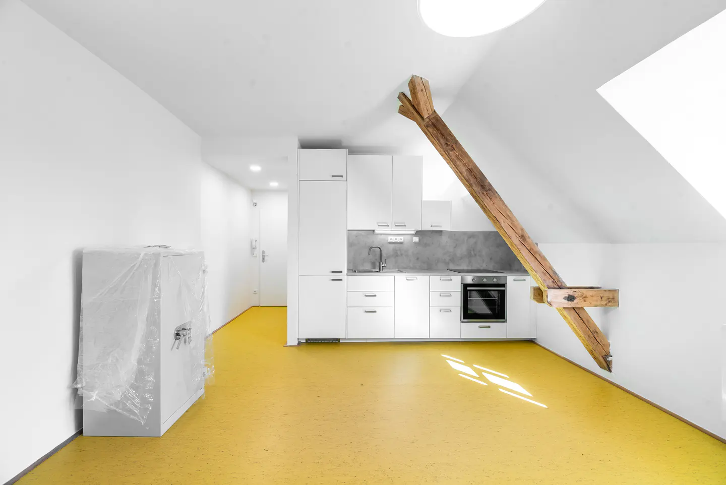 Bright, modern kitchen in an attic apartment with white cabinets, yellow floor, and exposed wooden beam. A wrapped appliance sits to the left.
