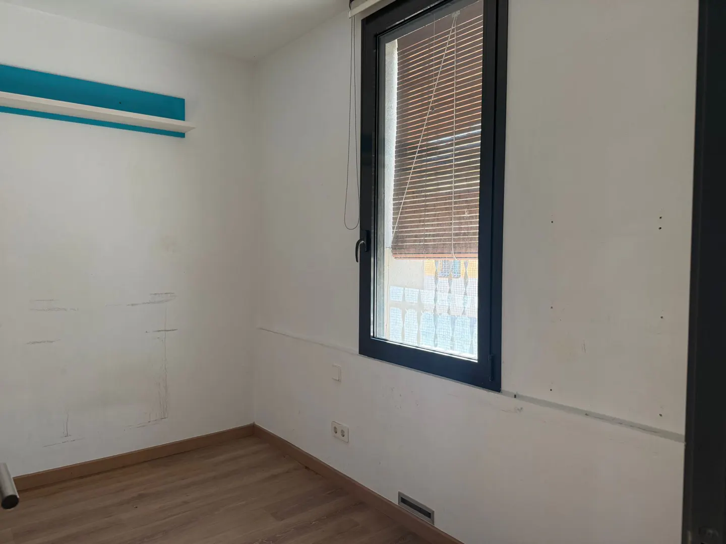 A bright, empty room with white walls, a wood floor, and a window with brown blinds. A blue accent stripe is near the ceiling.