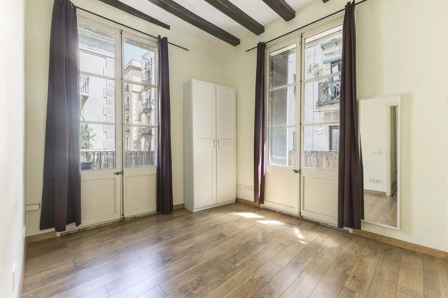 Bright room with wood floors, white walls, and exposed beams. Two windows with brown curtains flank a white wardrobe. A mirror hangs on the right wall.
