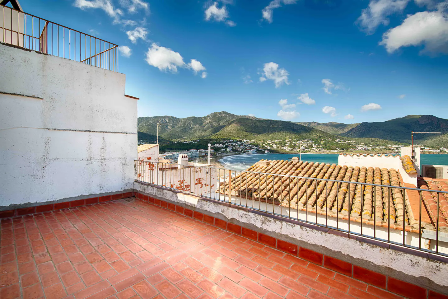 Terracotta-tiled rooftop terrace with metal railing overlooks a coastal town, beach, and mountains under a blue sky with scattered clouds.