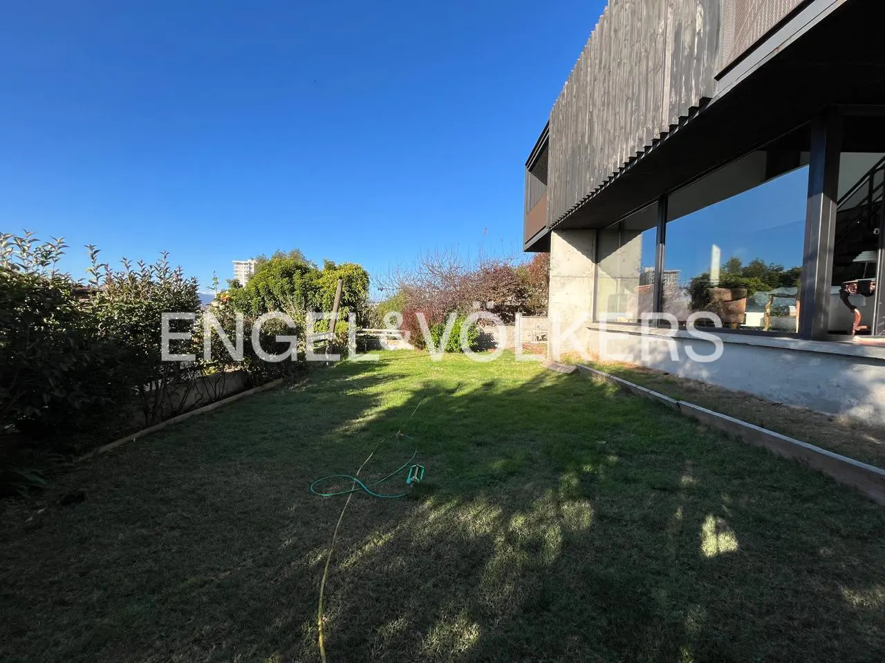 A modern home with a green lawn, trees, and blue sky. The Engel & Volkers logo is in the center of the image.