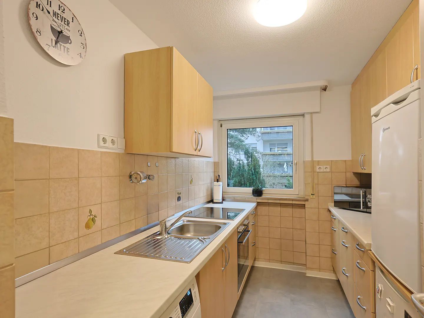 A bright kitchen with light wood cabinets, beige tile backsplash, and stainless steel sink. A window overlooks greenery. A clock hangs on the wall.