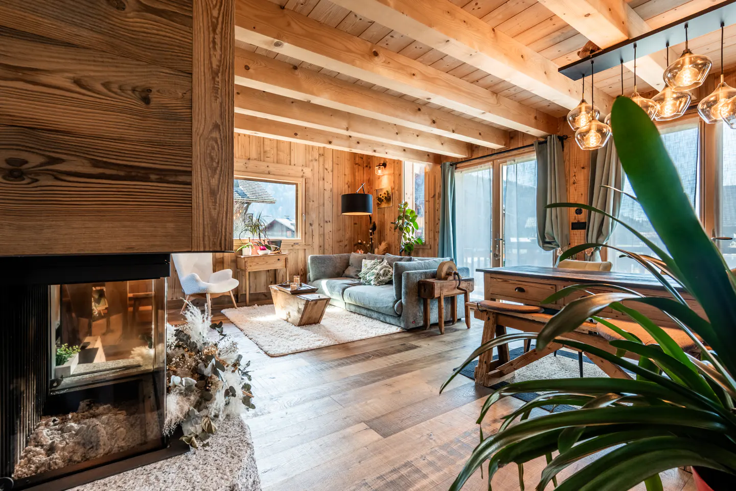 Living room with wood walls, beams, and floors. A gray sofa sits on a white rug. A fireplace and a large plant are also visible.
