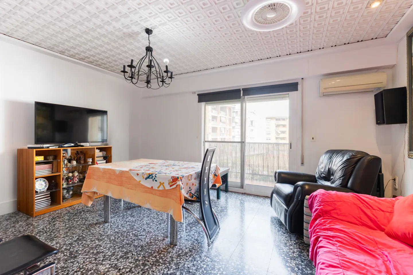 Living room with a TV, table, chairs, and a black leather armchair. The room has a patterned ceiling and a balcony with a view of the city.