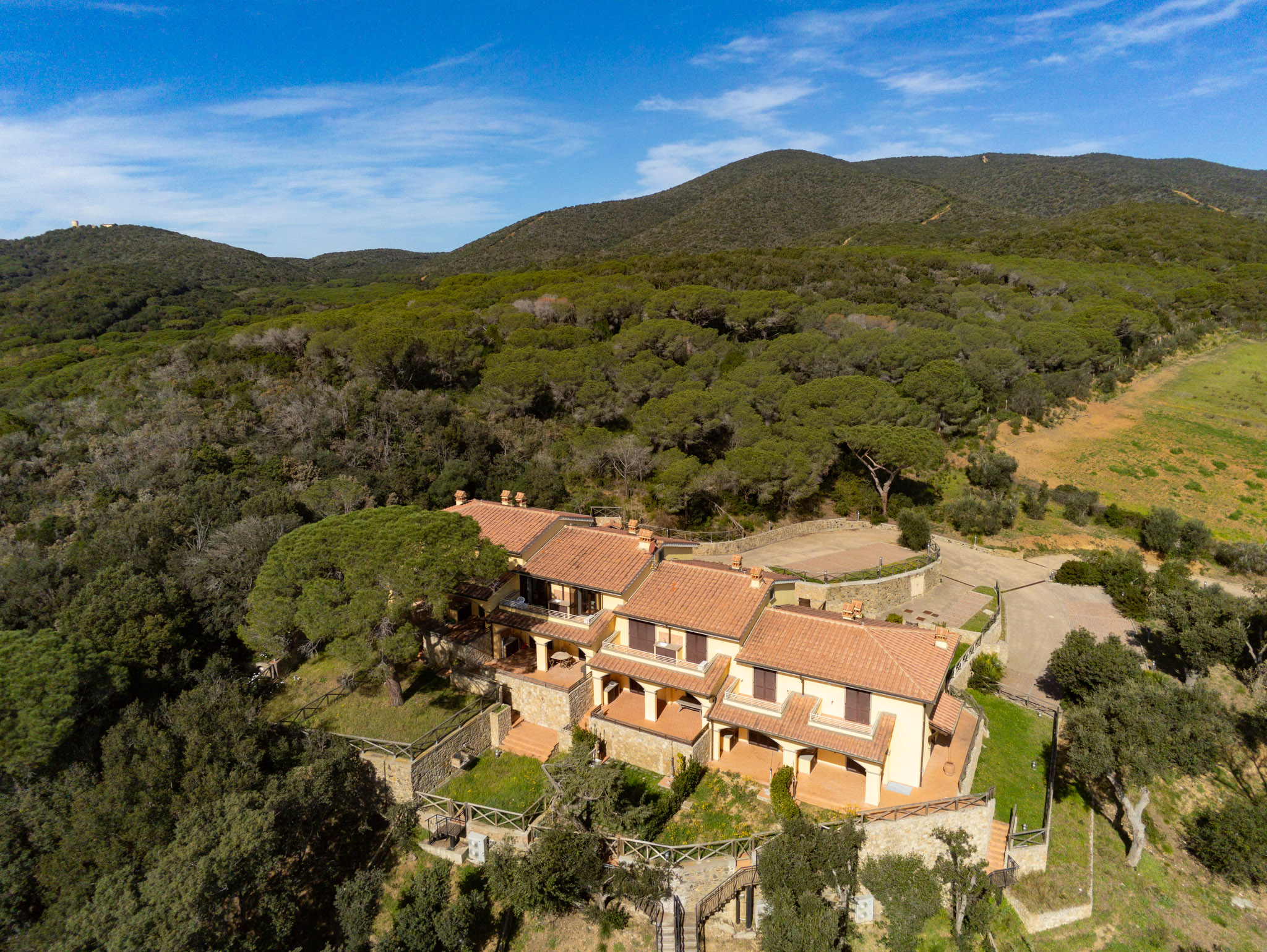 Aerial view of a yellow, multi-unit villa with a red tile roof, surrounded by green trees and hills under a blue sky.