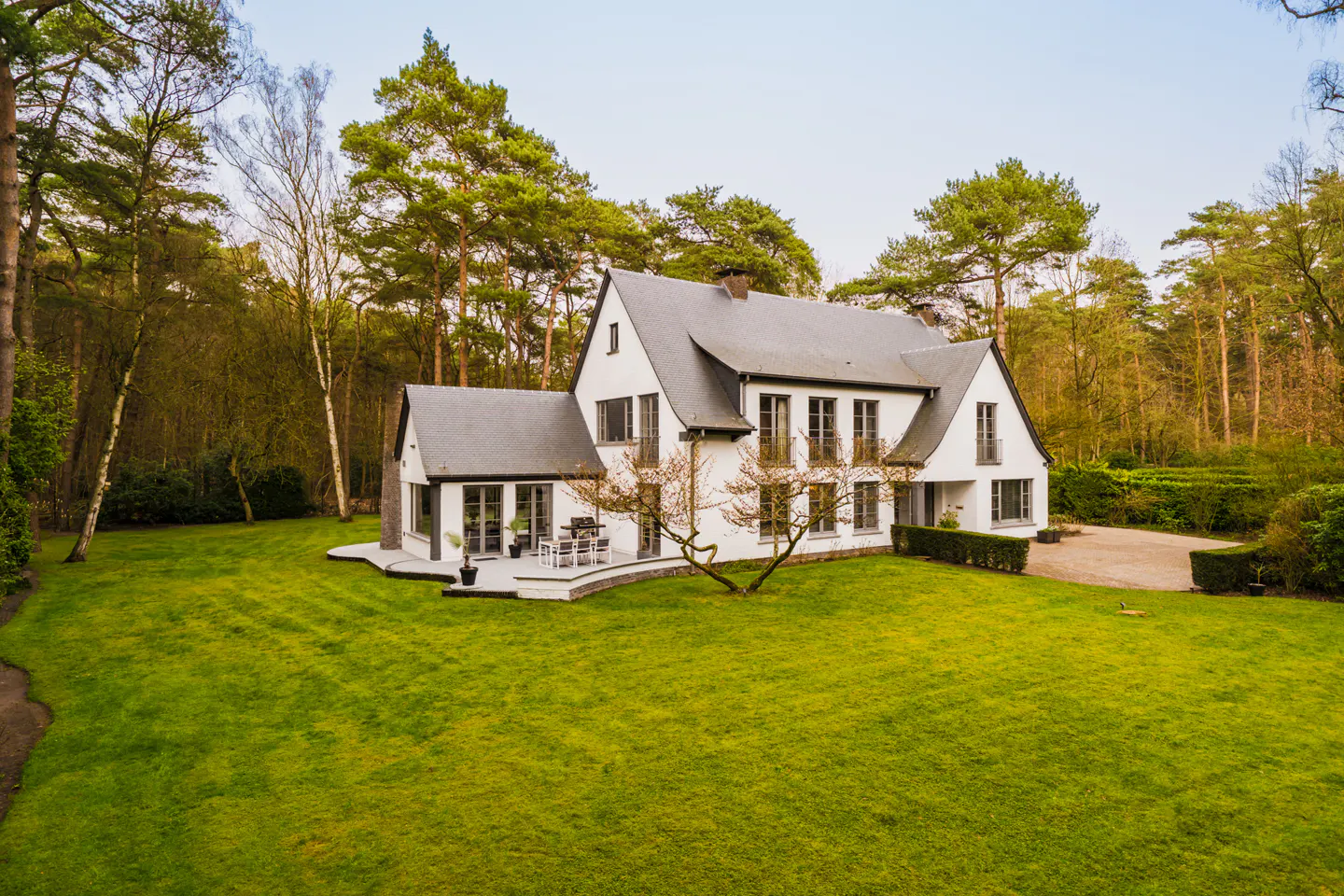 A white house with a gray roof sits on a large green lawn surrounded by trees.