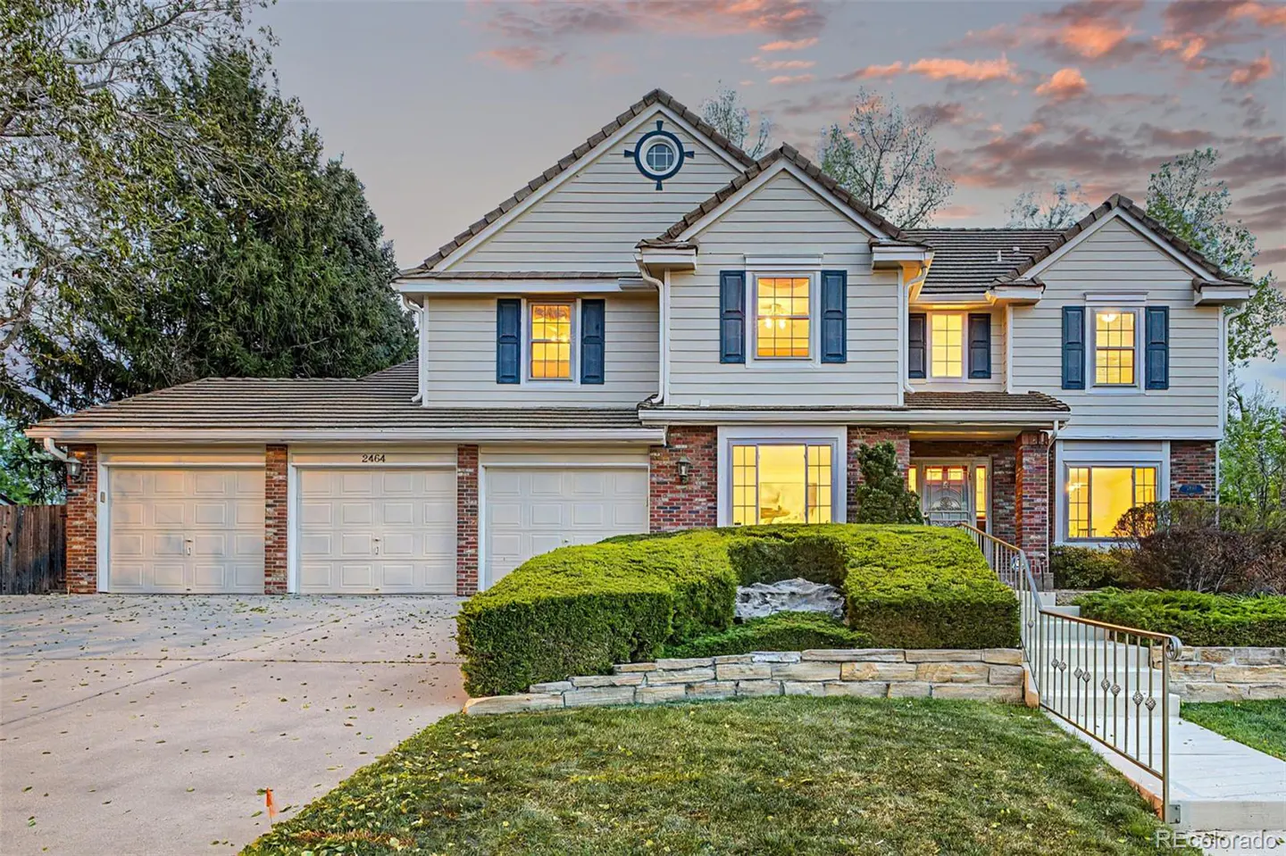 Two-story house with beige siding, brick accents, and blue shutters. A three-car garage is on the left, and manicured hedges line the front.