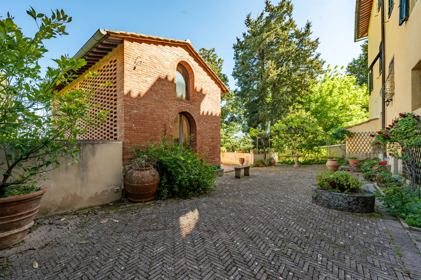 Brick building with arched windows and a lattice wall, surrounded by greenery and a herringbone brick patio.