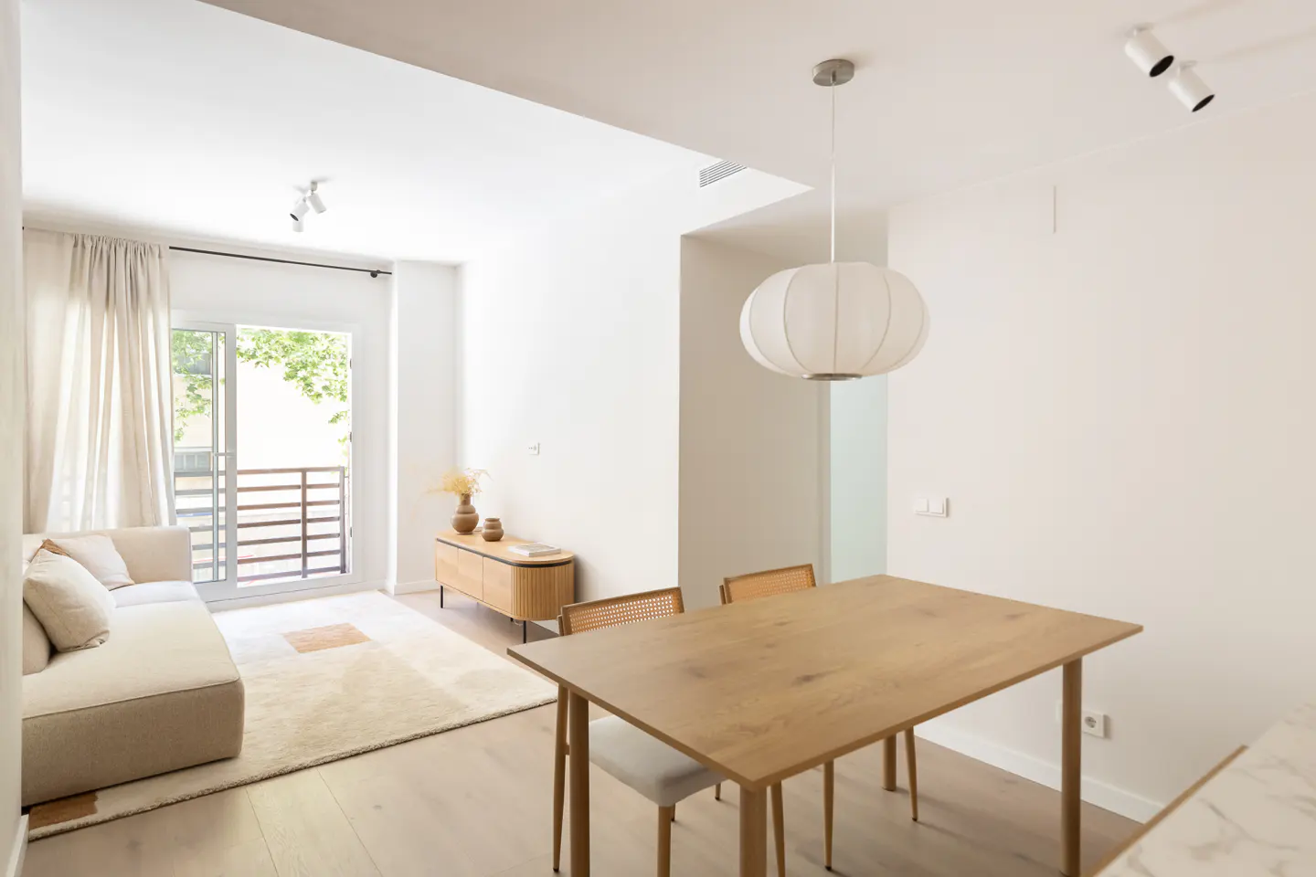 Bright, modern open-plan living and dining area with light wood furniture, a beige sofa, and a white pendant light.