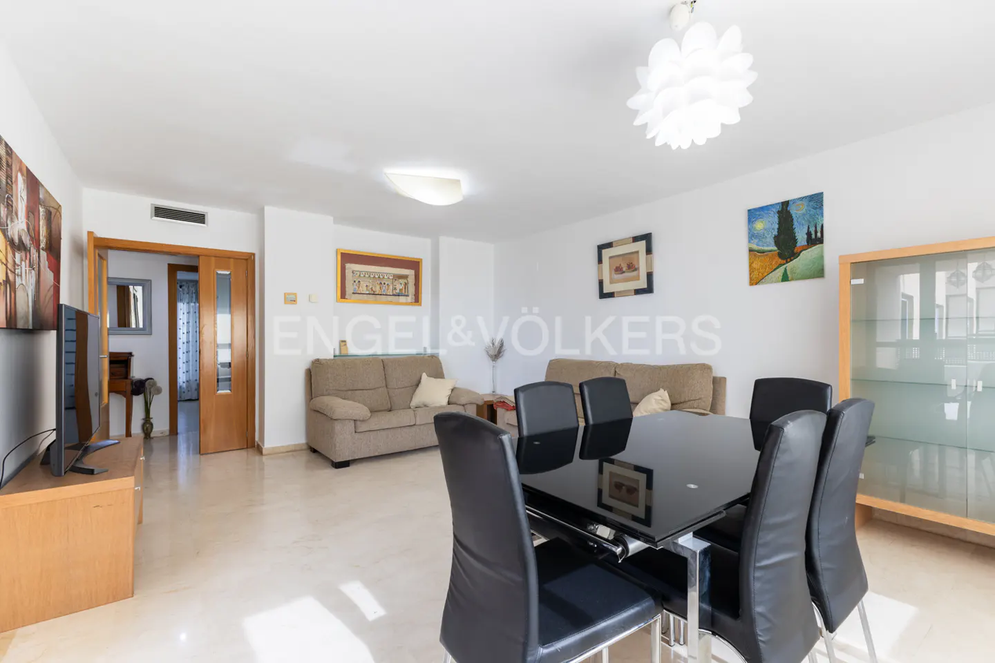 A bright living room with beige sofas, a black dining table with six chairs, and artwork on the white walls.