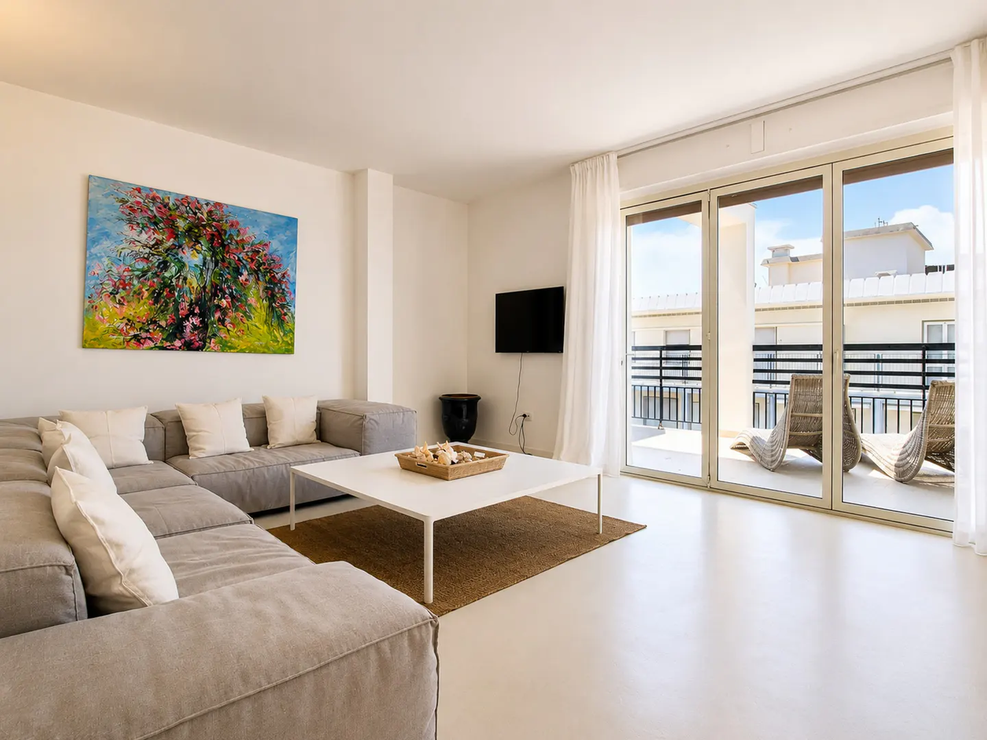 Bright living room with a gray sectional sofa, white coffee table, and a balcony with wicker chairs. A colorful painting hangs on the wall.