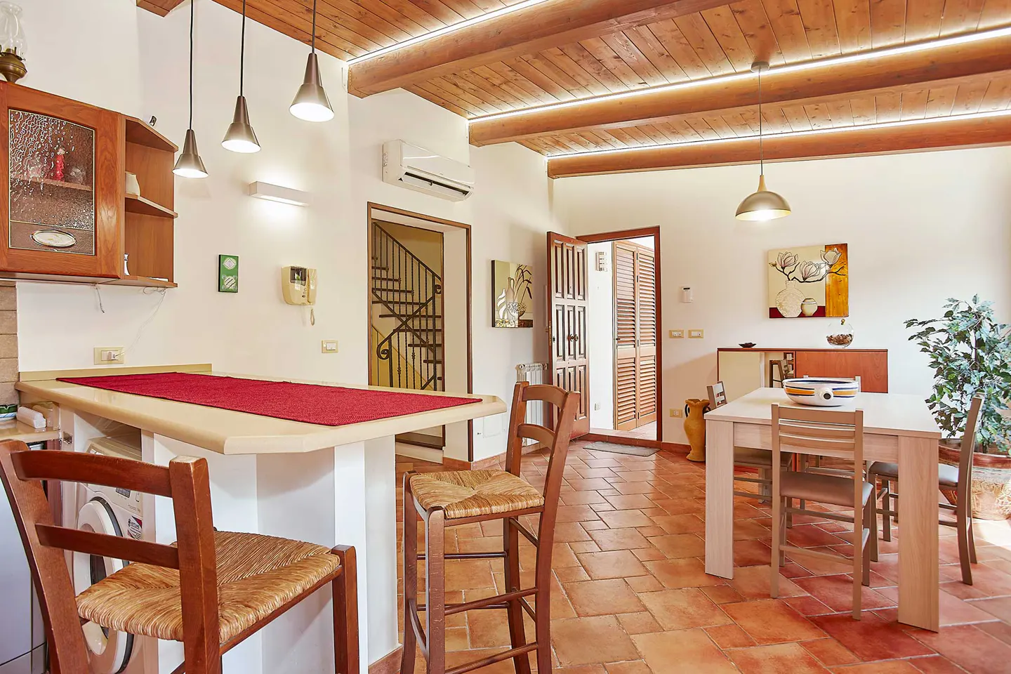 Interior view of a kitchen and dining area with terracotta tile flooring and a wooden beamed ceiling. A breakfast bar with stools is on the left, and a dining table is on the right.