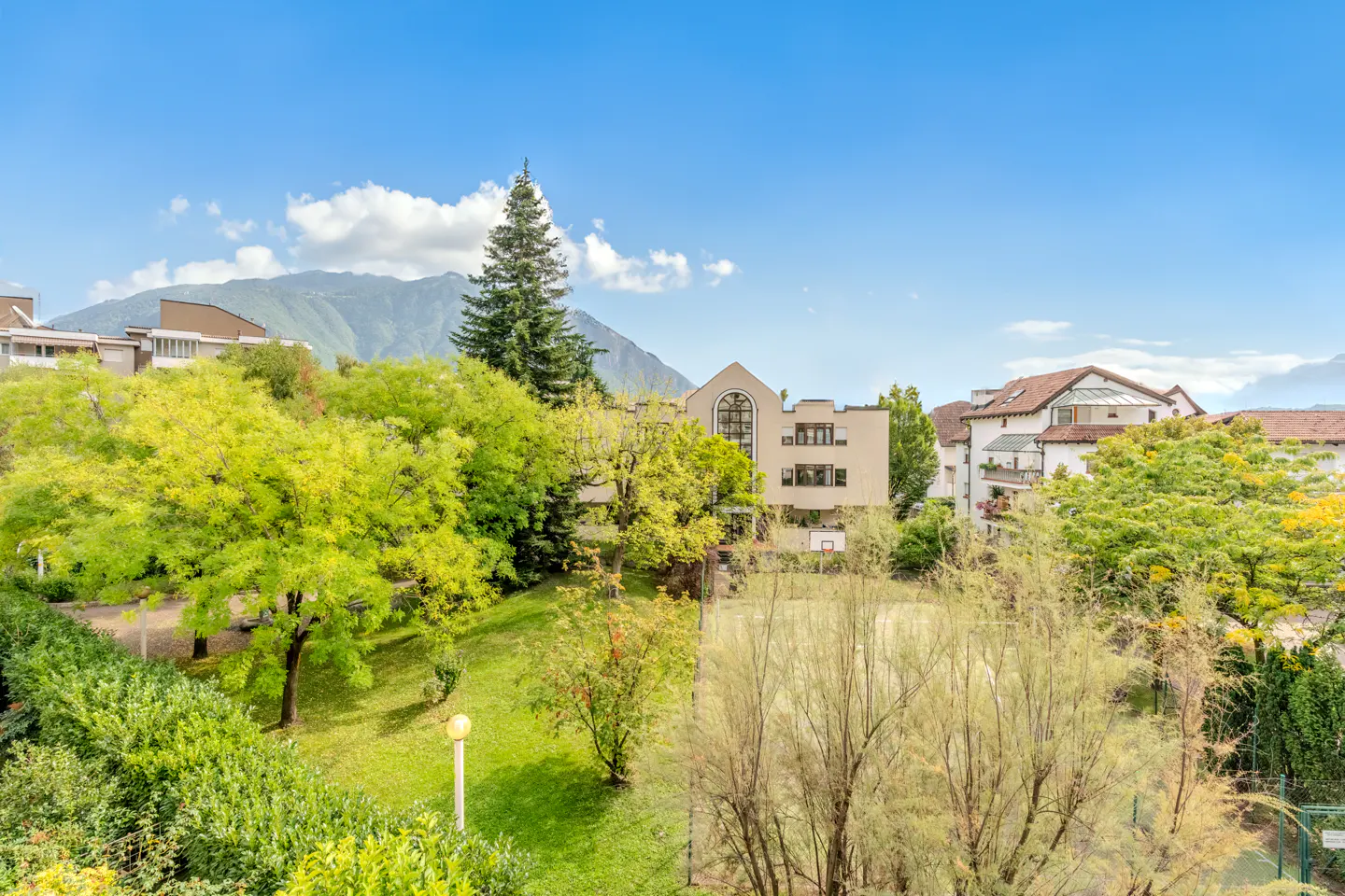 View of European town with green trees, buildings, and mountains under a blue sky.