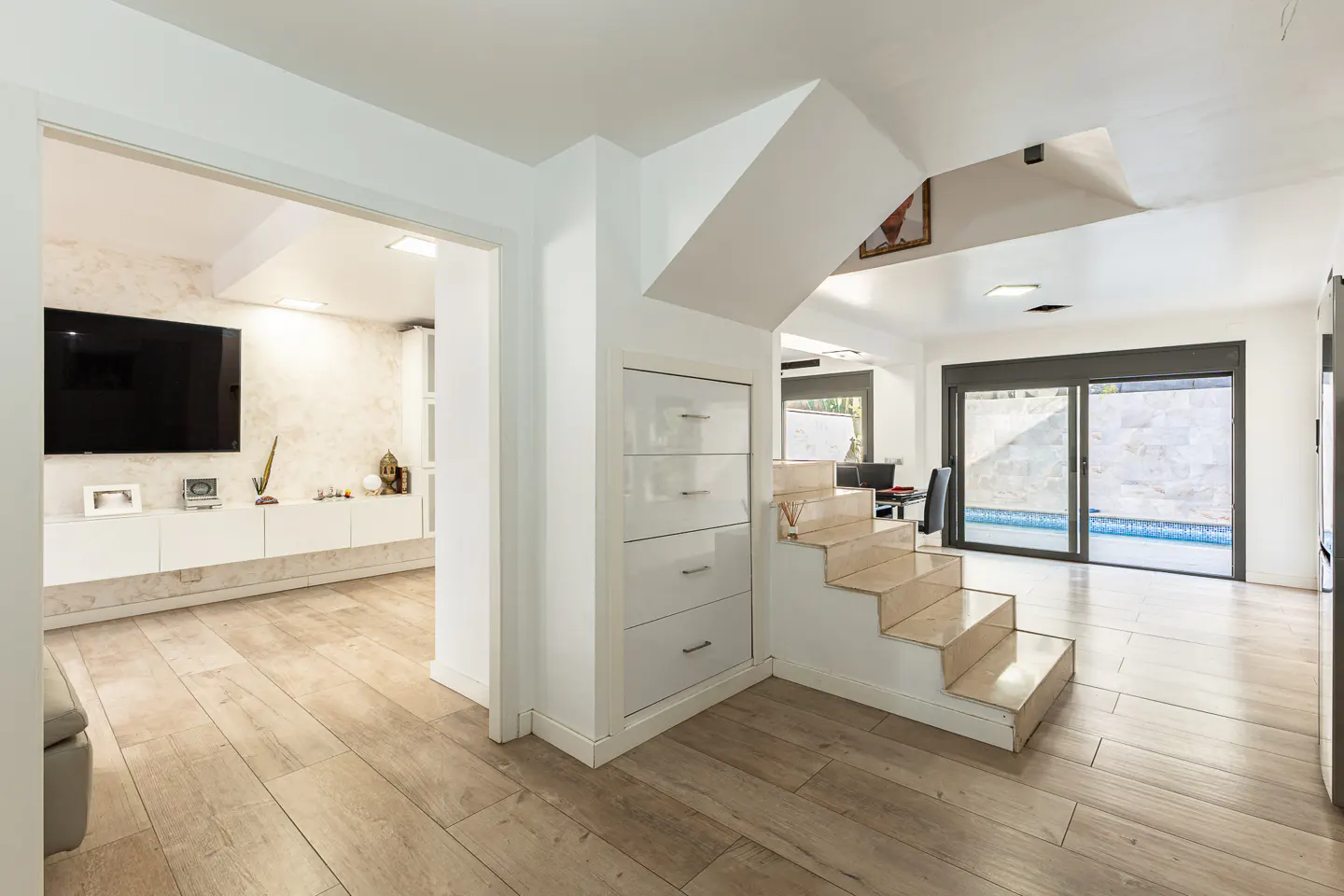 Bright, modern home interior with wood floors, stairs, and a view of a pool through sliding glass doors. A living room is visible through an open doorway.