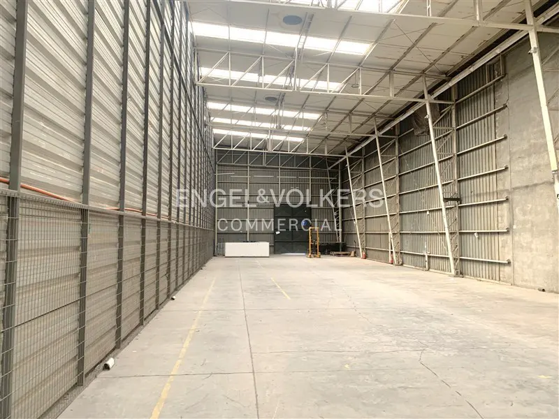 An empty warehouse with metal walls and a high ceiling with skylights. The floor is concrete and there is an Engel & Volkers logo in the center.