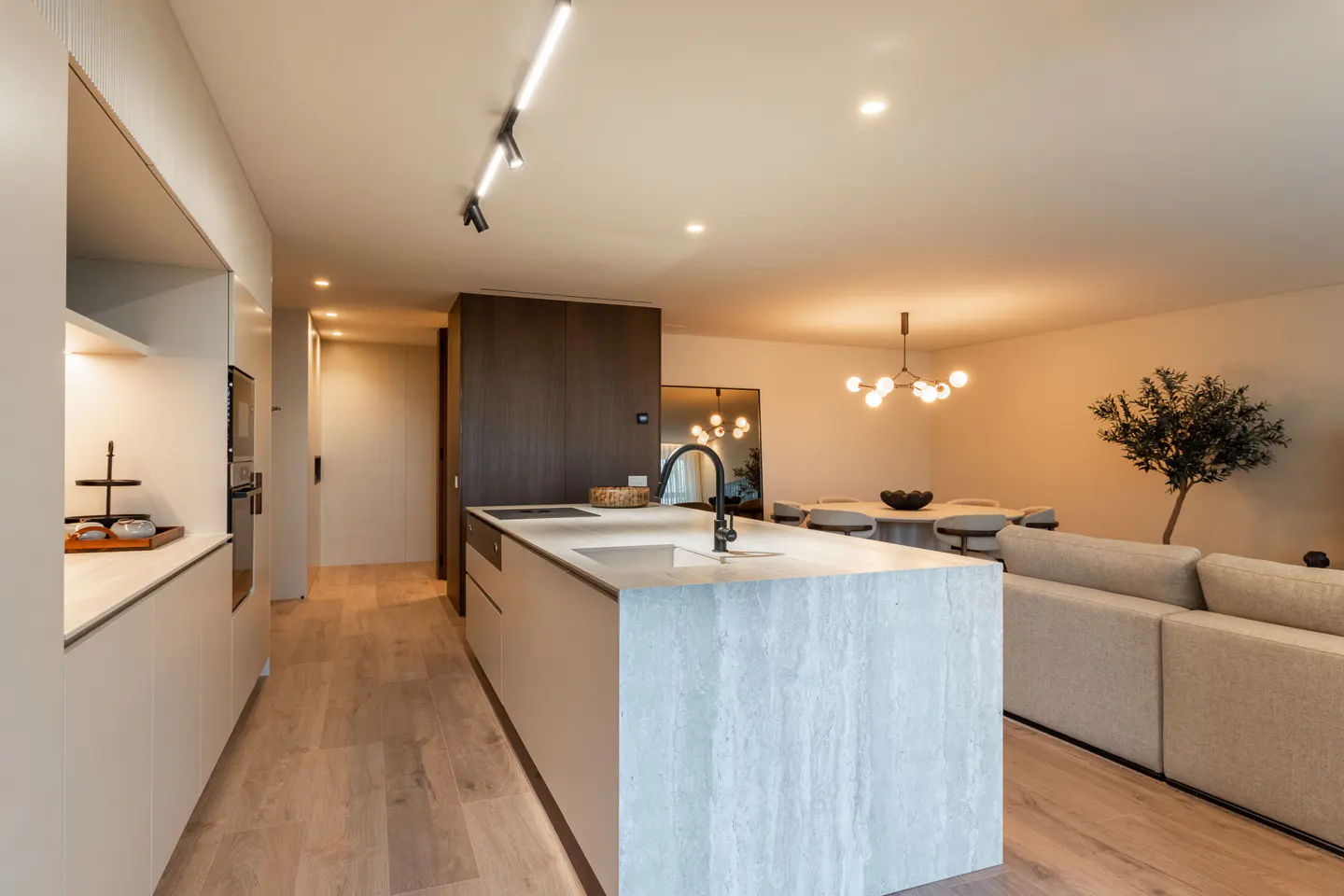 Modern open-plan kitchen with a stone island, light wood floors, and a dining area with a sofa in a neutral color palette.