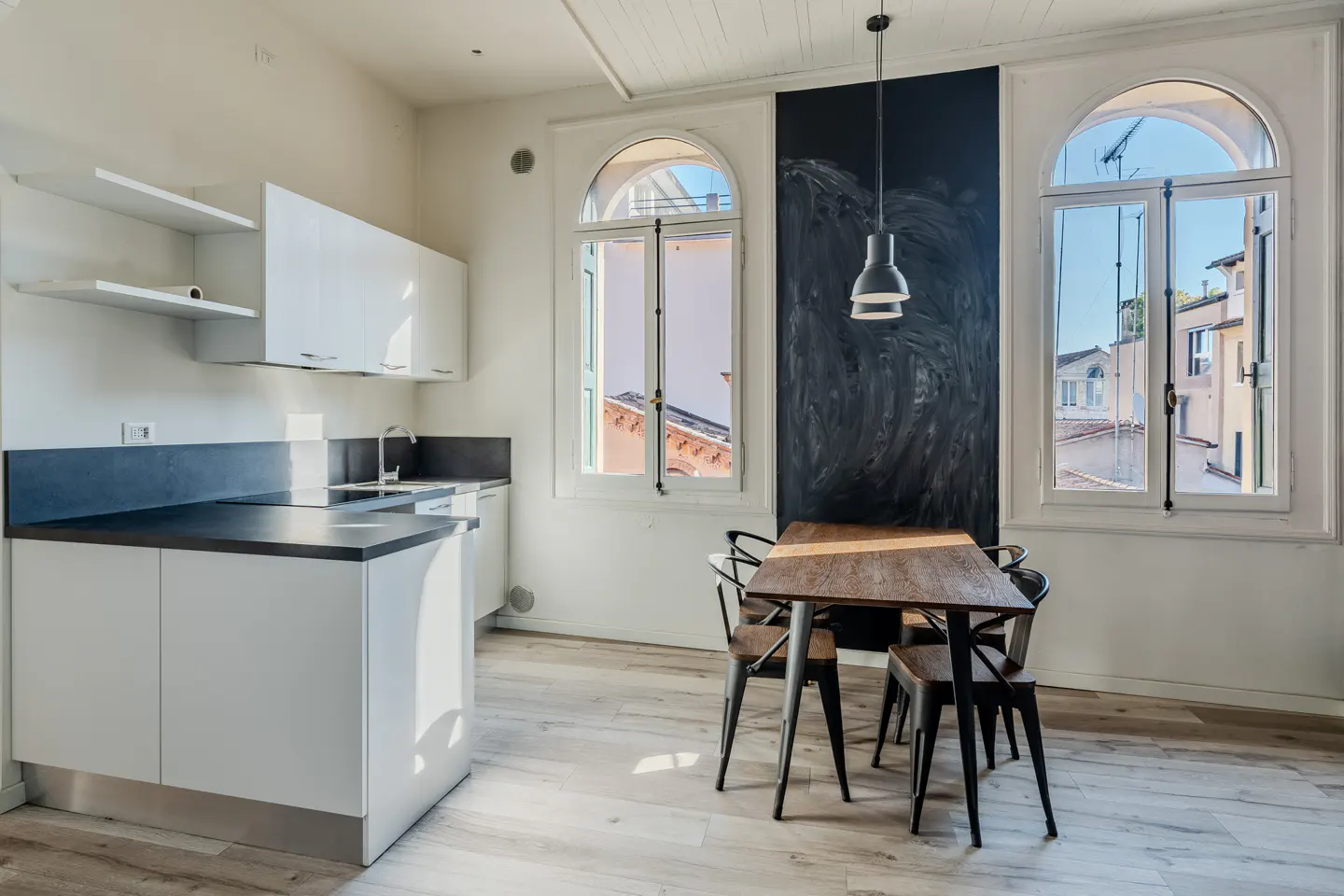 Bright kitchen with white cabinets, dark countertops, and a wooden table with four chairs near arched windows.