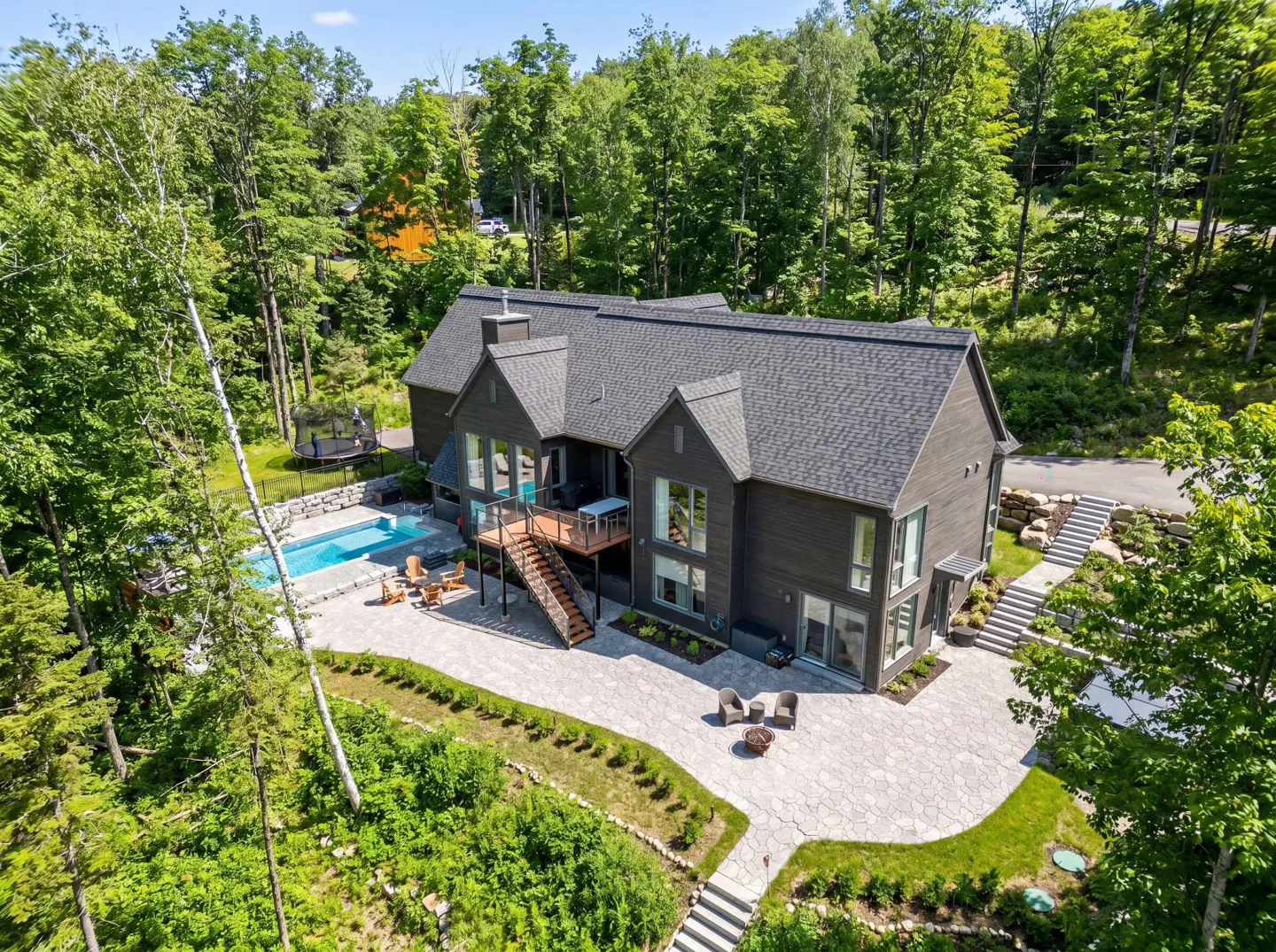 Aerial view of a modern, dark gray house with a pool, patio, and deck surrounded by lush green trees.