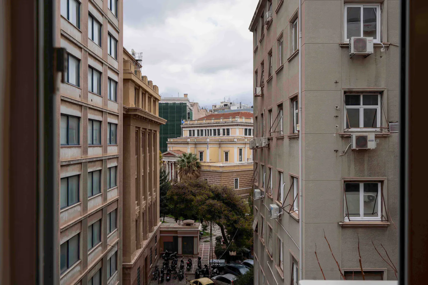 View from a window between buildings, showing a yellow building with columns and trees in the distance.