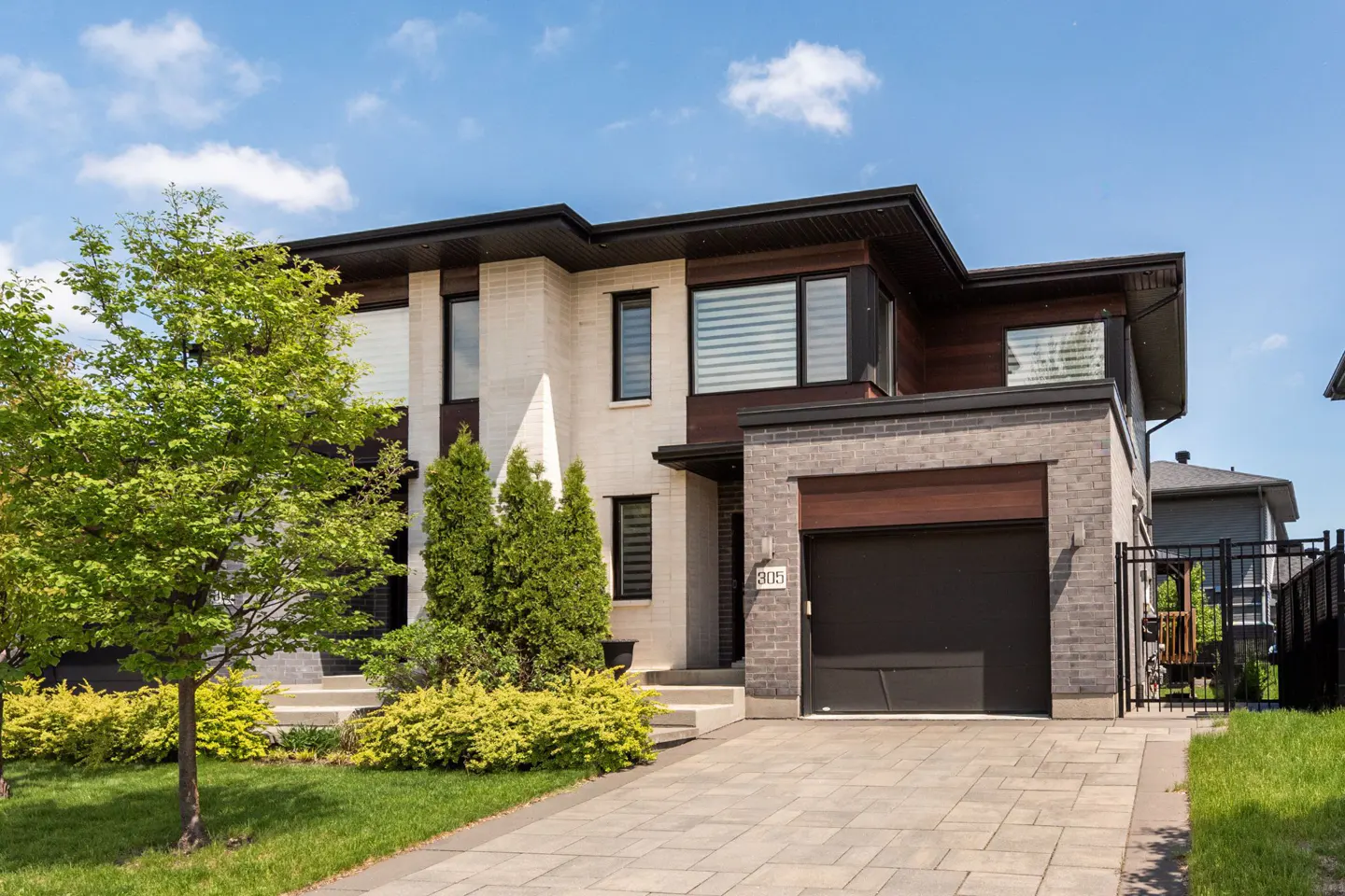 Modern two-story house with a dark garage door, brick and wood accents, and a paved driveway on a sunny day.