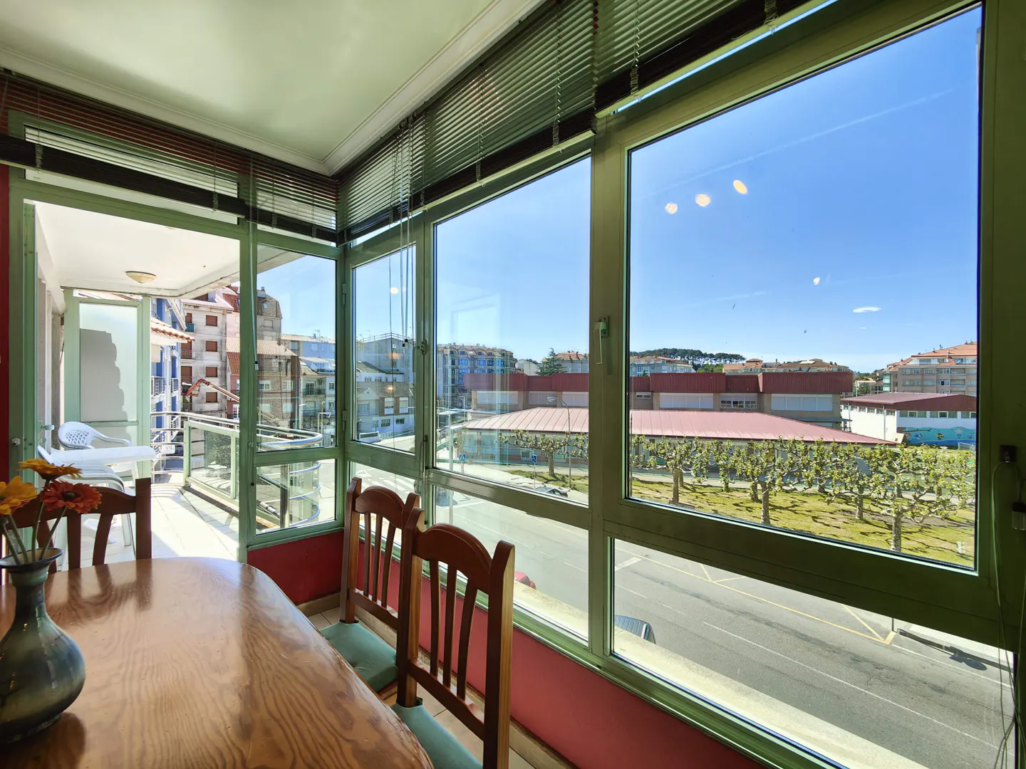 Bright dining room with wood table, chairs, and vase of flowers. Large windows offer a city view with blue sky. Balcony visible.