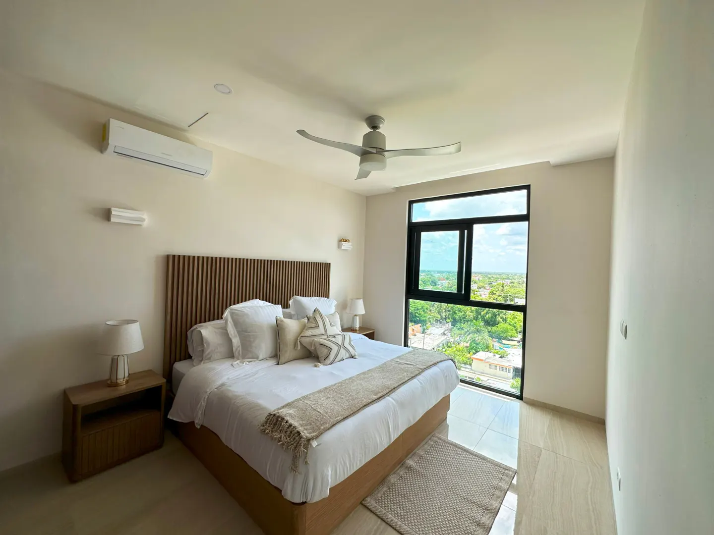 Bright bedroom with a wooden headboard, white linens, and a large window overlooking a green landscape. A ceiling fan and bedside tables are also visible.
