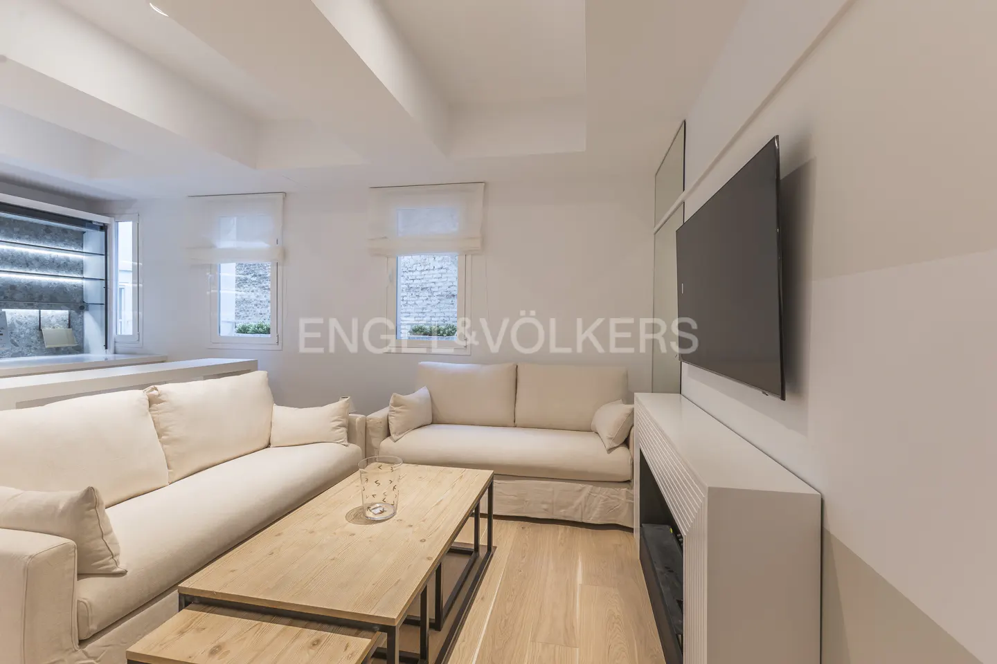 Living room with two cream sofas, a wooden coffee table, and a large TV mounted on the wall. Two windows with white blinds. Engel & Völkers logo in the background.