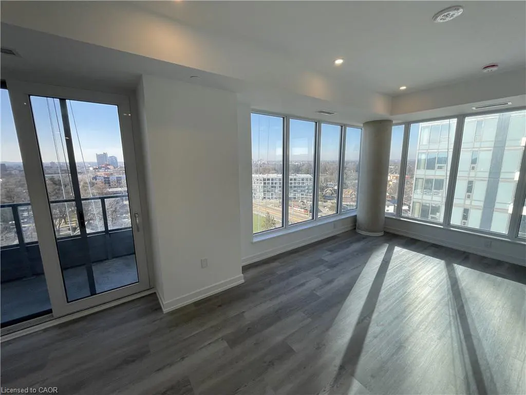 Bright, empty condo room with gray wood floors, white walls, and floor-to-ceiling windows showing a city view. A sliding glass door leads to a balcony.