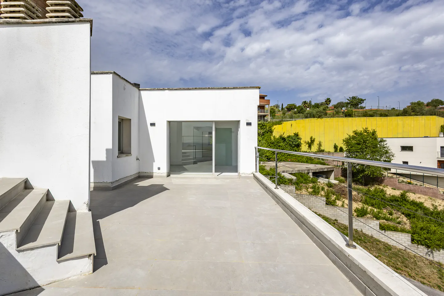 Wide shot of a modern white building with a large patio, sliding glass doors, and a metal railing overlooking greenery.