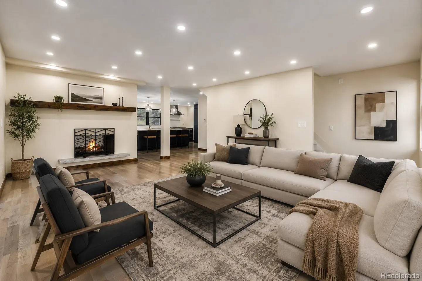 Living room with a fireplace, a beige sectional sofa, a dark wood coffee table, and two armchairs. The kitchen is visible in the background.