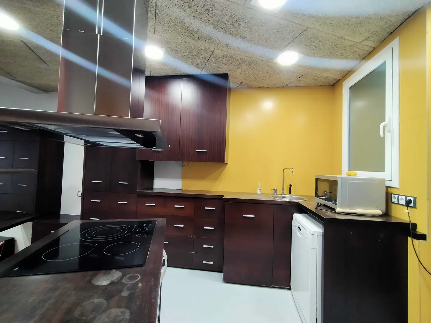 A kitchen with dark wood cabinets, a stainless steel range hood, and a yellow wall. A white window is on the right.