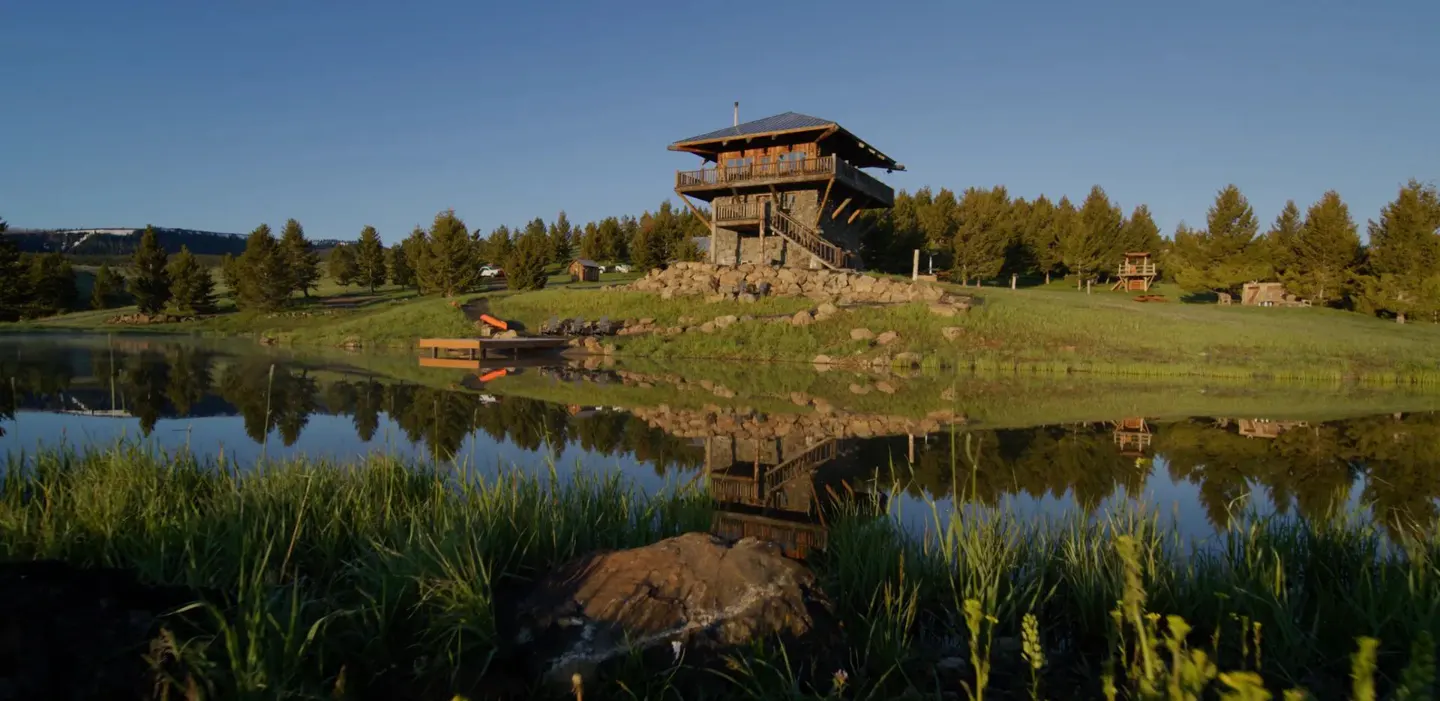 A stone and wood tower house sits on a hill overlooking a pond, reflecting in the water. Green grass and trees surround the property.