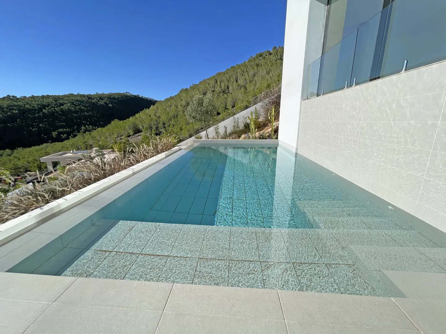 Modern infinity pool with light blue tiles overlooking a lush green hillside under a clear blue sky.